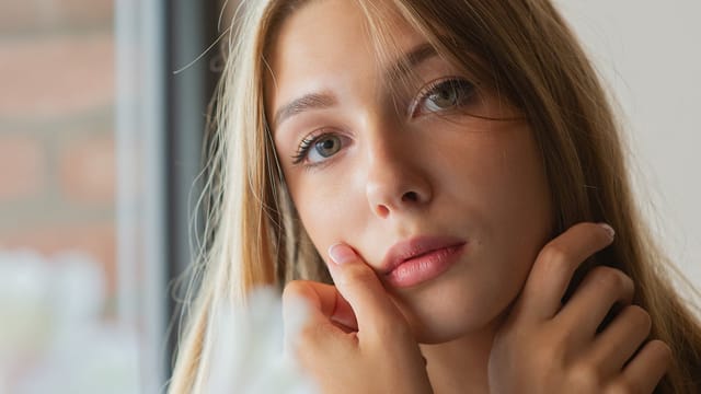 A young woman with long blonde hair gazes softly at the camera, her face framed by her hands. She has light eyes and a gentle expression, while natural light illuminates her features, subtly reflecting an air of hyper-independence.