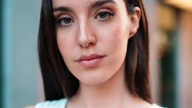 A young woman with long dark hair and brown eyes gazes into the camera with a neutral expression, subtly reflecting signs of unhappiness, as she wears a white top. The background is softly blurred, highlighting her face, quiet behaviors, and natural features.
