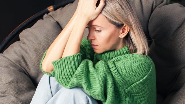A woman with blonde hair in a green sweater sits curled up on a chair, looking down with a distressed expression, her hand resting on her forehead.