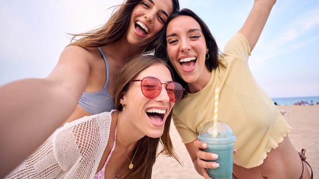 Three young women smiling and posing for a selfie on a sandy beach. One woman holds a blue drink with a striped straw. The sky is clear and they look happy, enjoying their time together outdoors.