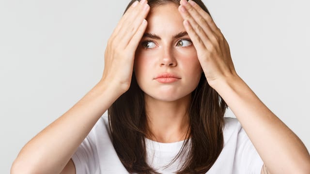 A woman with straight brown hair wearing a white shirt holds her hands to her forehead and looks to the side with a concerned or thoughtful expression against a plain light background.
