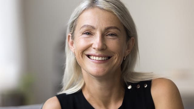 A middle-aged woman with long, straight, gray-blonde hair smiles warmly. She is wearing a sleeveless black top and small hoop earrings, and appears to be in a softly lit indoor setting.