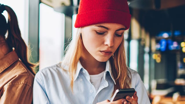 A young woman in a red beanie and light blue shirt looks intently at her smartphone while standing indoors near large windows.