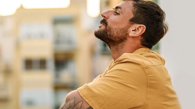 A man with short brown hair and a beard, wearing a mustard yellow shirt, sits outdoors with his arms crossed and eyes closed, appearing relaxed. The background is softly blurred with buildings and warm light.