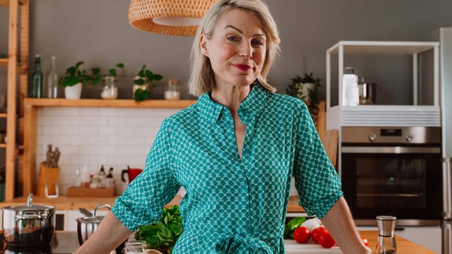 A smiling woman with short blonde hair wearing a teal patterned dress stands in a modern kitchen with wooden shelves, green plants, and fresh vegetables on the counter.