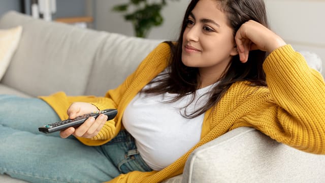 A young woman in a yellow sweater sits on a couch, smiling and relaxed, holding a TV remote control and looking toward a screen out of frame.