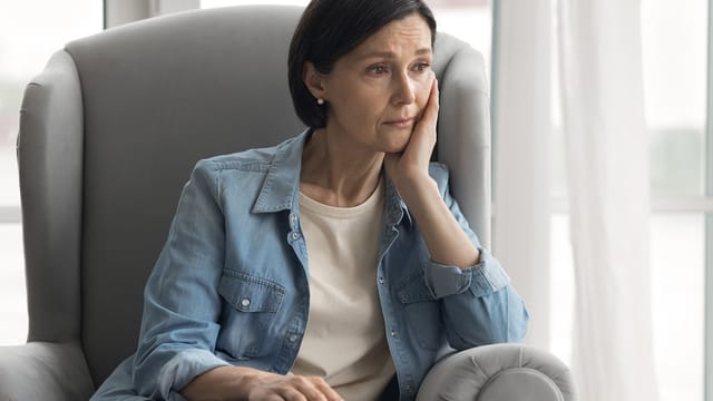 A woman with short dark hair sits in a gray armchair, resting her head on her hand and looking thoughtful or concerned. She wears a light denim shirt over a white top. Light streams in from a window behind her.