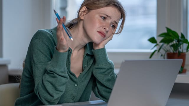 A woman sits at a desk in front of a laptop, looking confused and frustrated, with her head resting on one hand and holding a pen in the other. A plant is on the windowsill in the background.