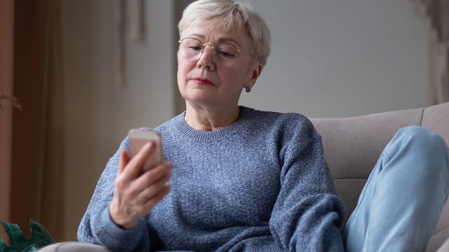 Older woman with short gray hair and glasses sits on a couch, wearing a blue sweater, looking thoughtfully at a smartphone in her hand.