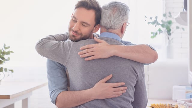 A younger man warmly hugs an older man with gray hair in a bright, cozy kitchen. Both appear content and comfortable, sharing a moment that beautifully reflects the bond of parenthood amid soft natural light and plants in the background.