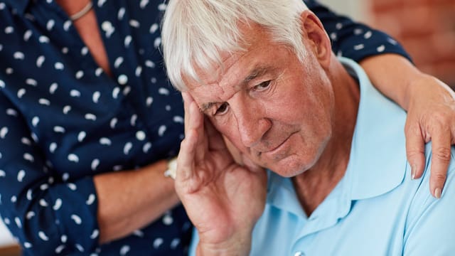 An elderly widower with gray hair looks distressed, resting his head on his hand. A person beside him in a blue patterned shirt gently touches his shoulder, offering comfort during a moment of loneliness and life after loss.