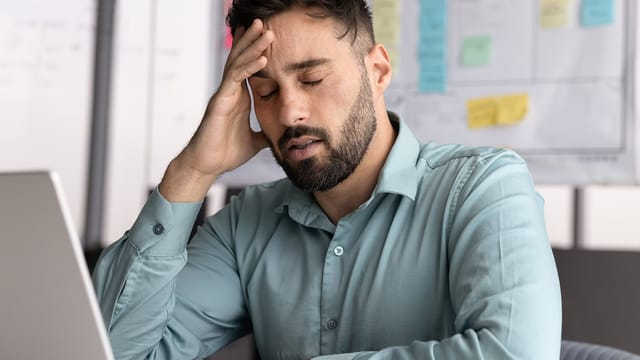 A man with a beard in a teal shirt sits at a desk, appearing stressed and employee overworked, with his eyes closed and hand on his forehead, as his laptop and whiteboard sit in the background.