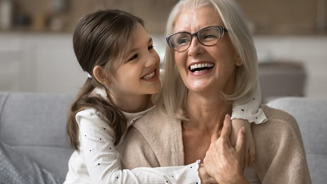 Smiling elderly woman with gray hair and glasses sits on a couch as her grandchild hugs her from behind; both look happy and joyful in a cozy home setting, reflecting the special bond between grandparents and their grandkids.