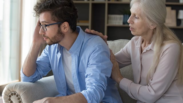 An older woman, likely his parent, sits beside a younger man on a couch, gently touching his shoulder and back as he looks stressed. Their serious conversation hints at navigating toxic patterns in adulthood.