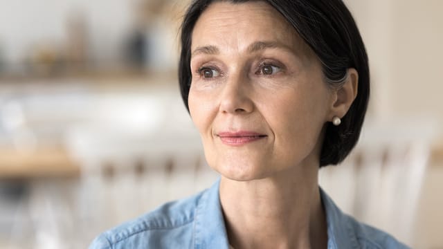 A middle-aged woman with short dark hair and pearl earrings looks thoughtfully into the distance, perhaps reflecting on generational parenting, wearing a light blue shirt with a softly blurred indoor background.