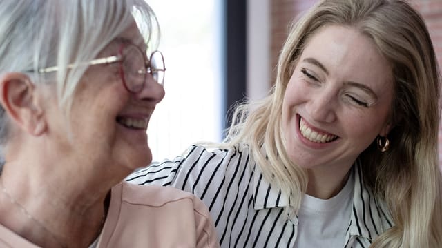 A young woman with long blonde hair smiles warmly at an older woman with gray hair and glasses. Both are laughing, enjoying each other's company—a beautiful moment of intergenerational friendship.