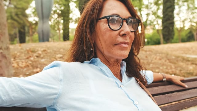 A woman with long brown hair and glasses sits on a bench outdoors, wearing a light blue shirt. She looks to the side with a thoughtful expression, perhaps reflecting on self-worth, surrounded by trees and greenery.