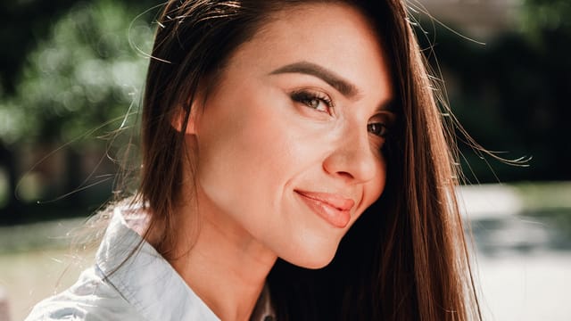 A woman with long brown hair and natural makeup smiles softly at the camera outdoors, surrounded by greenery and sunlight, radiating calm as she enjoys a peaceful moment of coping with burnout.