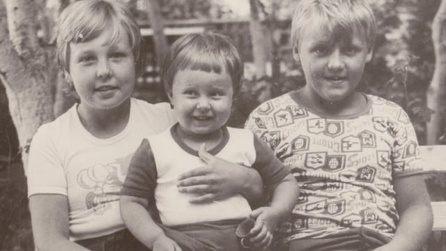 Three children sit closely together on a bench outdoors. The child in the middle, a toddler, smiles while being held by the older children on either side—a charming black and white image reflecting generational differences.