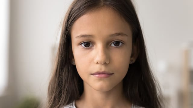 A young girl with long brown hair and brown eyes, shaped by quiet traits, looks directly at the camera with a neutral expression. She is indoors, with a softly blurred background hinting at early brushes with adulthood.