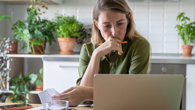 A woman, raised by a single parent, sits at a kitchen table, looking thoughtfully at a laptop screen while holding a piece of paper and an orange pen. Potted plants and kitchen items are visible in the background.