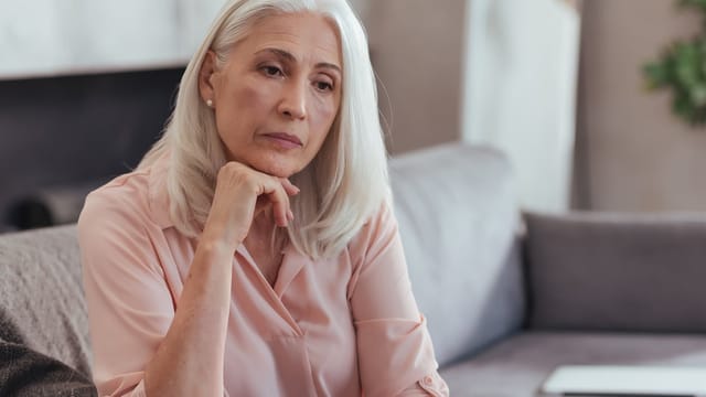 An older woman with long gray hair sits on a couch, resting her chin on her hand and looking thoughtful or concerned—perhaps reflecting on parent child relationships or the psychology after 60. She wears a light pink blouse and appears deep in thought.