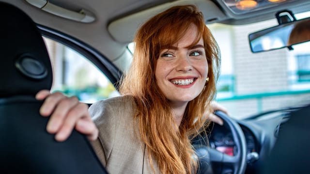 A woman with long red hair smiles while sitting in the driver’s seat of a car, looking back over her shoulder. The car interior is visible, and she appears cheerful and confident, perhaps reflecting positive parking habits.