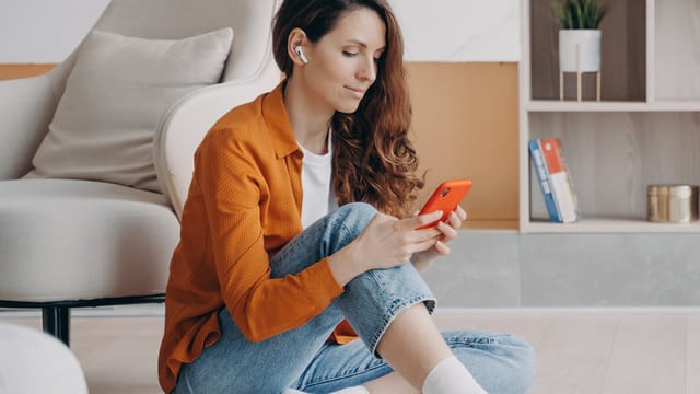 A woman with long brown hair sits on the floor, wearing wireless earbuds that provide background noise. Dressed in an orange shirt and jeans, she holds a red smartphone and looks at it, alone with her thoughts. Shelves and a chair are visible in the background.