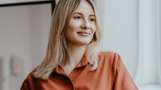 A woman with straight blonde hair and a nose ring, wearing an orange shirt, smiles softly while looking to the side in a bright indoor setting, capturing a rare moment of peace for her usually restless mind.