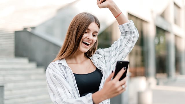 A young woman with long brown hair smiles and raises her arm in excitement while looking at her smartphone outdoors, energized by social energy as she stands near concrete steps and modern buildings.