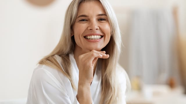 A smiling woman with long blonde hair, feeling like herself, wears a white blouse and rests her chin on her hand. She is indoors, with a soft, neutral background out of focus.