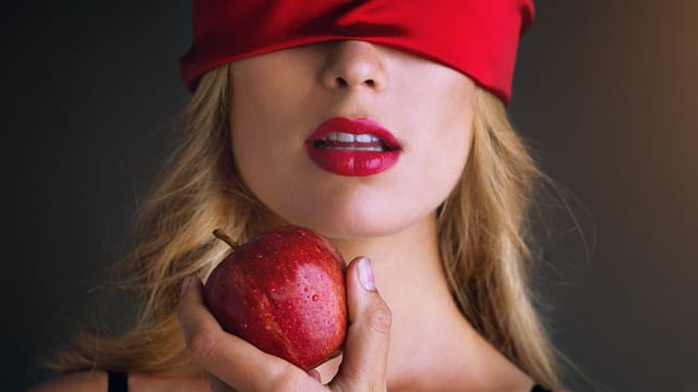 A woman with blonde hair and red lips wears a red blindfold, holding a shiny red apple close to her mouth. The dark, blurred background sets the mood of romantic temptation.