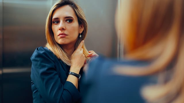 A woman with blond hair and a navy blue top looks at her reflection in a mirror, adjusting her hair with a thoughtful expression, contemplating her imaginary self—a moment echoing the psychology of unhealthy comparison.