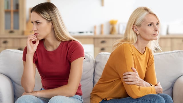 Two women sit on a couch facing away from each other, both looking upset and avoiding eye contact. The younger woman in a red shirt appears thoughtful, while the older adult in a mustard sweater has her arms crossed, reflecting tense psychology.
