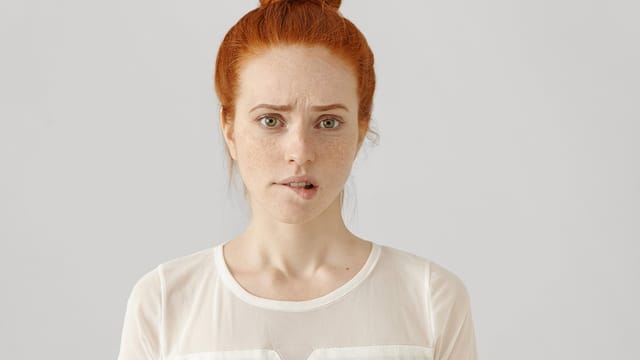A young woman with red hair in a bun, wearing a sheer white top, looks directly at the camera with a concerned expression, raising one eyebrow and biting her lower lip, as if contemplating whether to apologize or start taking the blame against a plain light background.