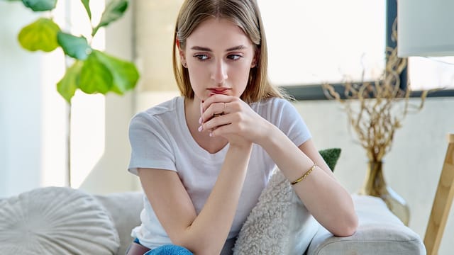 A young woman with long hair sits on a couch indoors, resting her chin on her hands and looking thoughtful, perhaps reflecting on childhood habits. She wears a white t-shirt and blue jeans, with a window and plant in the background.