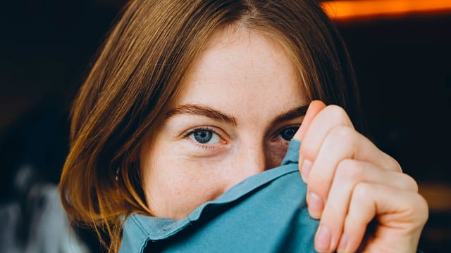 A person with light brown hair and blue eyes gazes into the camera, holding up a blue fabric that partially covers their mouth and nose—a subtle nod to the spotlight effect studied by psychologists, where we often overestimate how much others notice us.
