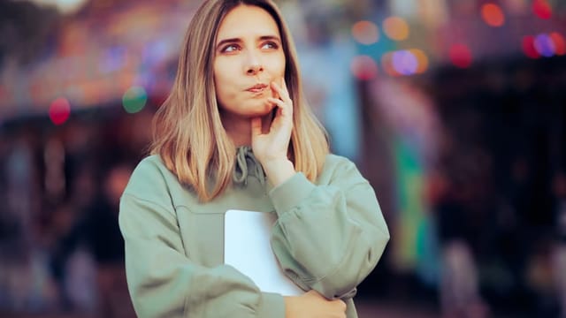 A young woman with light brown hair in a casual green sweatshirt holds a notebook and looks thoughtful, touching her lips with one finger. Colorful blurred lights glow in the background.