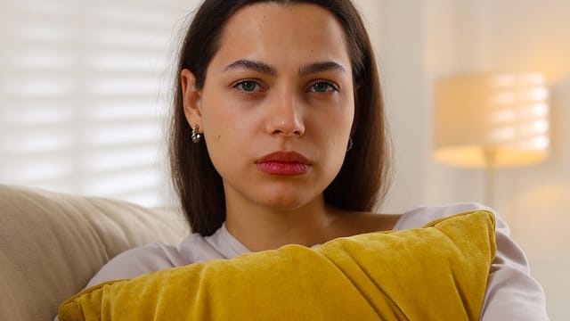 A woman with long brown hair, the eldest daughter, sits on a couch holding a yellow pillow. She has a neutral, slightly serious expression. A lamp and white blinds are visible in the softly lit background.