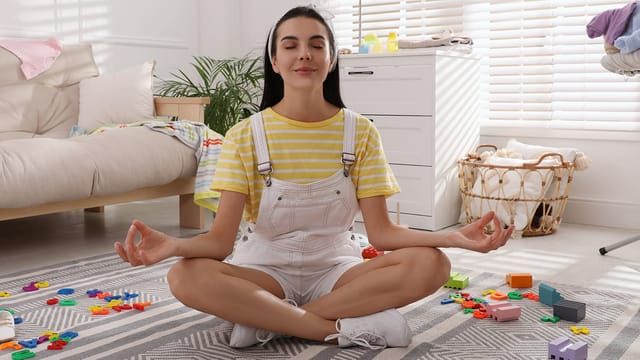 A woman in a striped yellow shirt and white overalls sits cross-legged on a rug, meditating with eyes closed, surrounded by colorful plastic toys in a bright living room that reflects real family dynamics and a happily lived-in home.