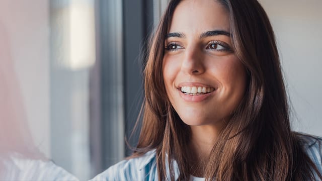 A young woman with long brown hair smiles while looking out of a window. She is wearing a light blue and white shirt, and natural light brightens her face, reflecting a hopeful expression amid emotional chaos.