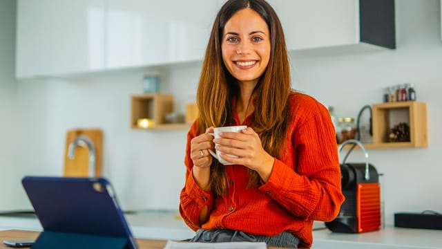 A woman with long brown hair, wearing an orange shirt, sits in a modern kitchen holding a white mug and smiling at the camera. A laptop and coffee machine are visible beside her as she enjoys her daily habits for productivity.