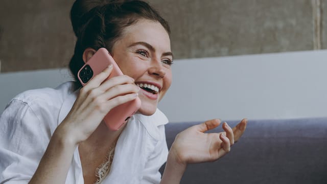 A smiling woman in a white shirt talks on her phone, holding it to her ear with one hand while gesturing with the other, sitting indoors on a sofa. Her warm expression suggests she's nurturing a close relationship, always being kind and attentive during conversations.