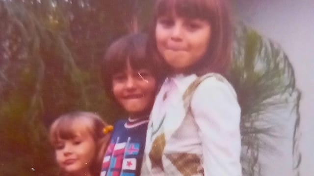 Three young children stand close together outdoors, smiling at the camera with a sense of confidence. The child on the right wears a white outfit, the middle child has a dark shirt, and the child on the left has blonde hair with bangs—a classic scene reminiscent of 70s kids. Greenery is visible behind them.