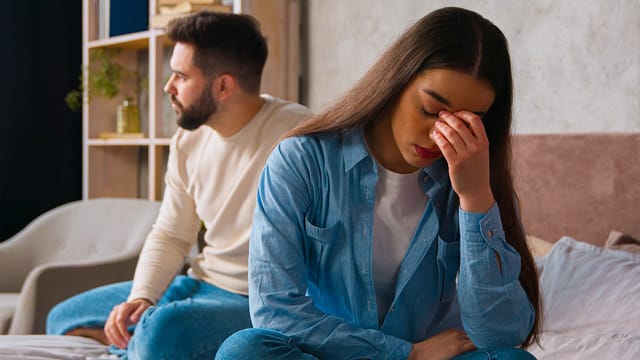 A woman sits on a bed, looking upset with her hand on her face, while a man sits behind her, turned away and gazing into the distance. Both appear distressed, suggesting a tense or emotional moment that may reflect challenges in personal growth or difficulties related to social habits.