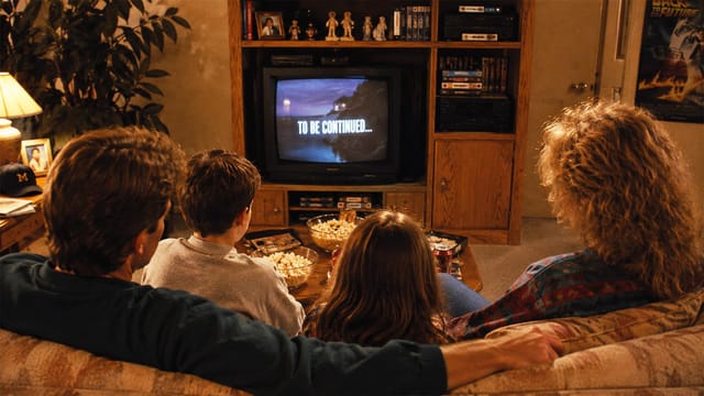 A family of four sits on a couch watching a TV that displays TO BE CONTINUED. They have popcorn bowls on the table, and the living room is warmly lit with shelves, movies, and decorations in the background.