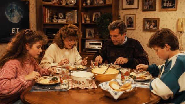 A family of four sits at a wooden table eating dinner together. The table has plates of food, bowls, and glasses. The room has framed photos, a bookshelf, and an E.T. movie poster on the wall.