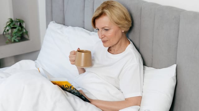 An older woman in her 70s with short blonde hair sits up in bed, enjoying her morning routines. She holds a mug and reads a book, dressed in a white t-shirt and covered by a white blanket. A small plant adds warmth to the background.