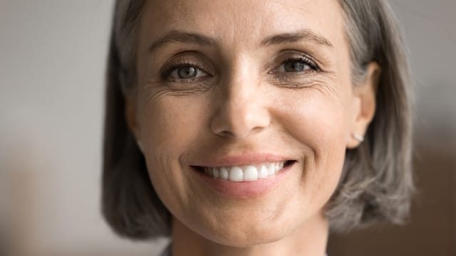 Close-up of a middle-aged woman with short gray hair, smiling warmly. Her eyes are bright, and she has subtle wrinkles, suggesting a genuine, happy expression. The background is softly blurred.