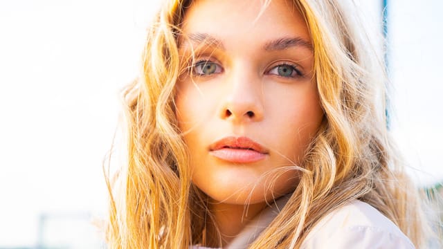 Close-up of a young woman with long, wavy blonde hair and blue-green eyes, looking directly at the camera with a neutral expression, in natural outdoor light.